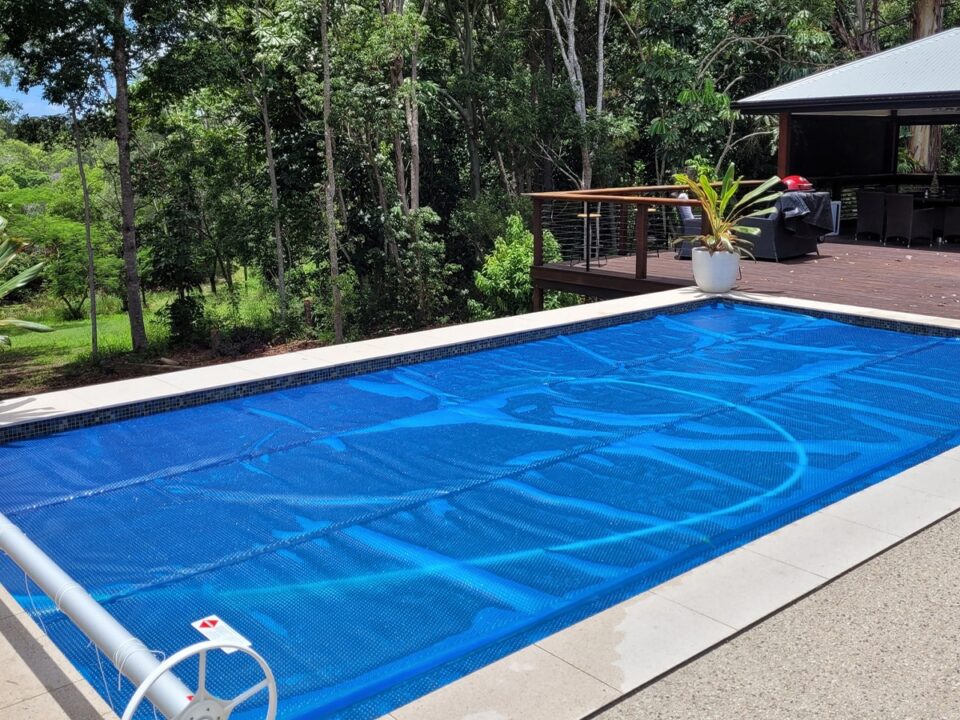 A backyard swimming pool winterised with a blue pool cover, surrounded by trees, with a wooden deck and gazebo in the background. | AES Pool Heating Solutions