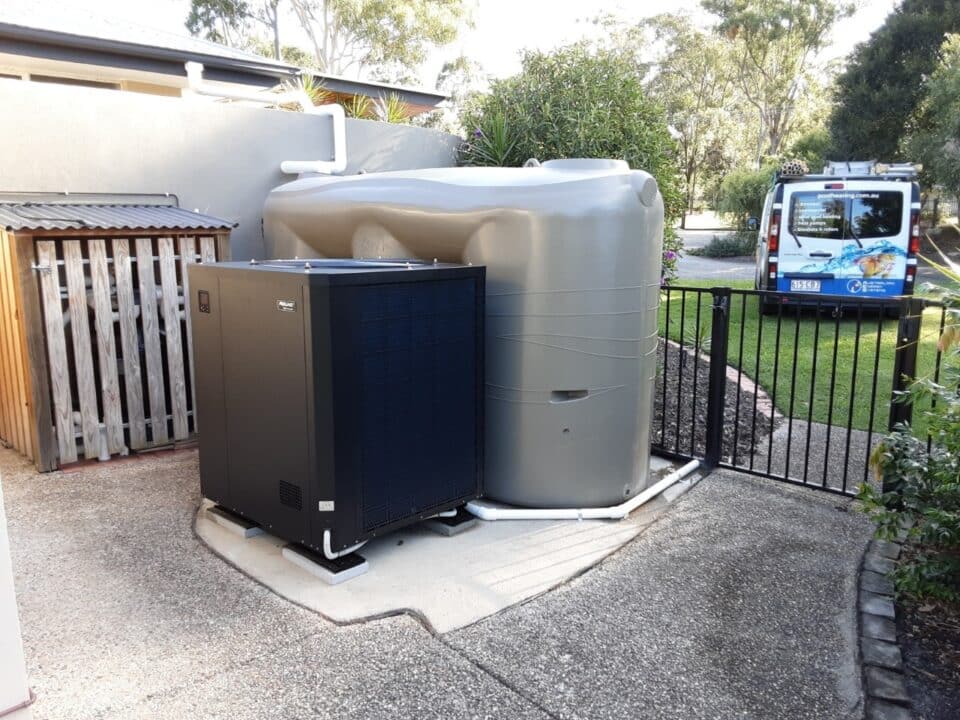 An outdoor setup featuring a large gray water tank connected to white pipes and a black heat pump installed on a concrete slab. The area is fenced with a black metal gate, and a service van is parked in the background near a green lawn and trees. | AES Pool Heating Solutions