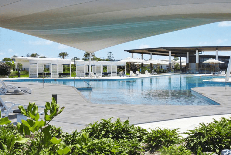 A luxurious outdoor pool area with modern lounge chairs, cabanas, and sun umbrellas. The pool is surrounded by greenery and open space, with a large canopy providing shaded areas. Enjoy the serene atmosphere at RV Lifestyle Village Oceanside, crafted for ultimate comfort under a clear blue sky. | AES Pool Heating Solutions