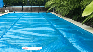 Blue solar pool cover on a tropical backyard pool with roller and palm trees